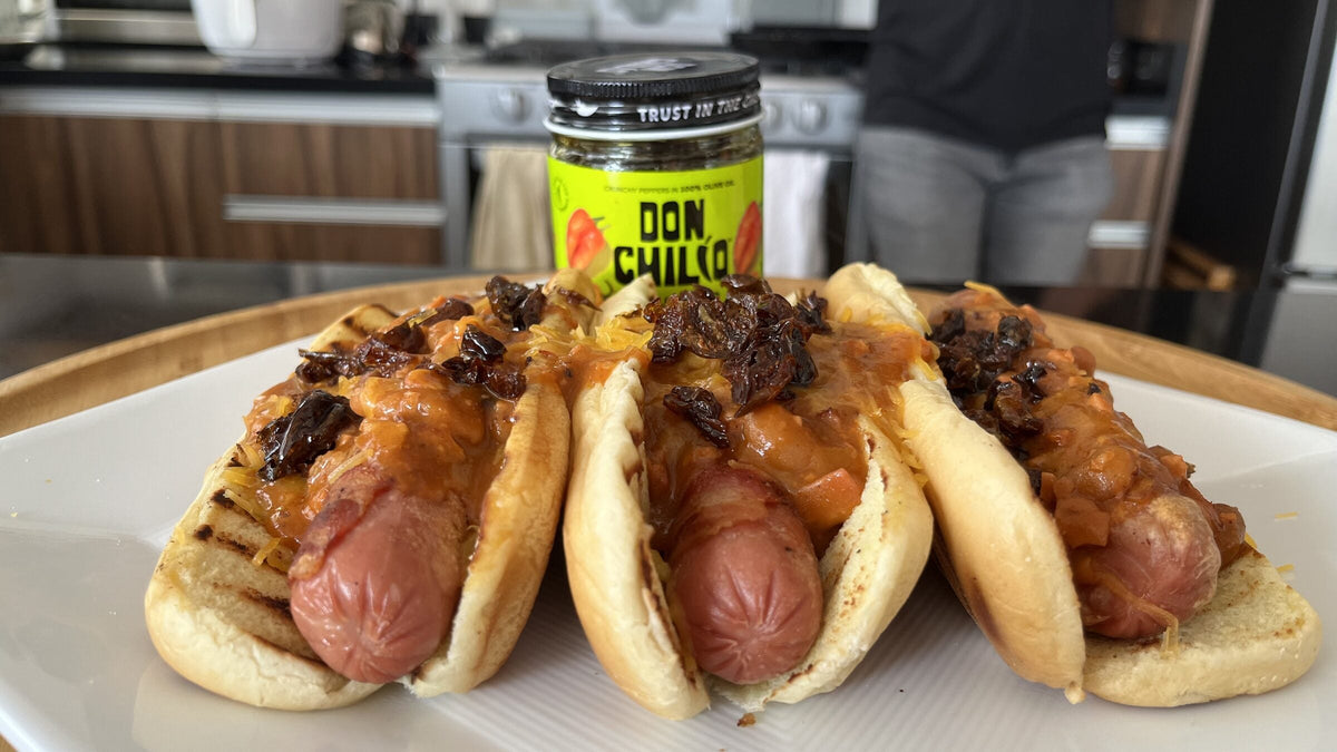 Three bacon-wrapped chili dogs topped with cheddar and Don Chilio Flamin' Habanero Chili Crisp, with the jar behind them