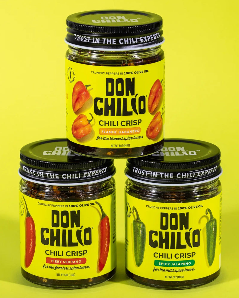 Three glass jars of Don Chilko Chili Crisp arranged in a pyramid on a bright yellow background. The top jar is labeled Flamin' Habanero, and the two bottom jars are labeled Firey Serrano and Spicy Jalapeño.