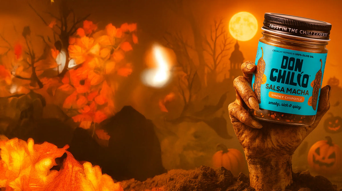 Zombie hand rising from the earth holding a jar of Don Chilio Smoky Chipotle Salsa Macha under a full moon, with jack-o'-lanterns